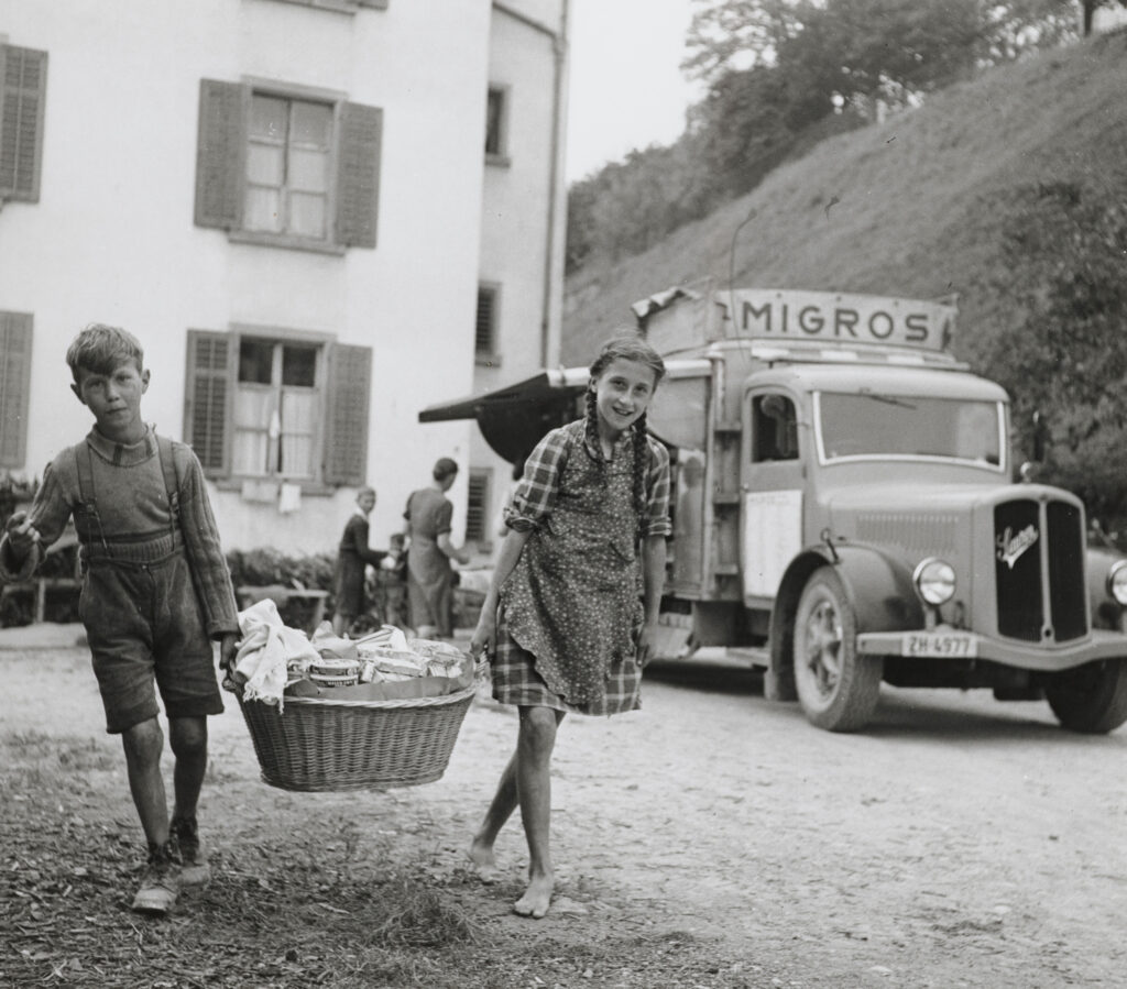 1925 tritt die Migros mit einer Niedrigpreisstrategie in den Lebensmittelmarkt ein. Von Quartier zu Quartier ziehende Verkaufswagen führen zu Beginn nur sechs Artikel im Sortiment. Diese sind dafür bis zu 25% günstiger als üblich. Migros-Verkaufswagen im Tösstal, Theo Frey, Weiningen, 1942. © Theo Frey / Fotostiftung Schweiz