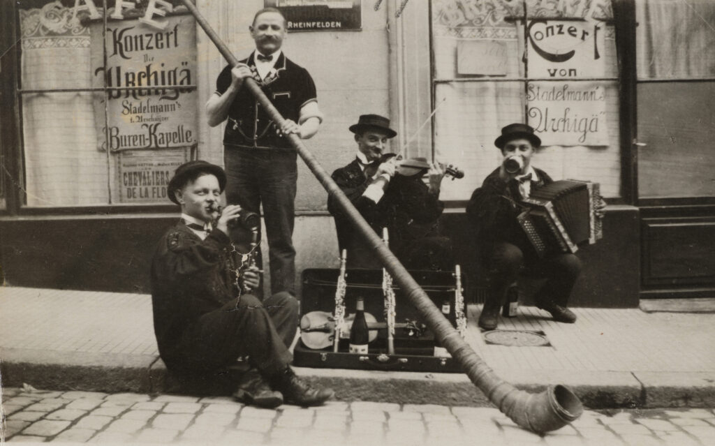 Kasi Geisser (links) und weitere Musiker posieren mit ihren Instrumenten und alkoholischen Getränken vor ihrem Konzertplakat. Die Innerschweizer vermitteln damit eine Garantie für feucht-fröhliche Tanznächte im Zürcher Niederdorf. Kasi Geisser (Klarinette); unbekannt (Alphorn); Emil Christen (Geige) und Roman Stadelmann (Handorgel) posieren vor Konzertplakat «Urchigä», Zürich, um 1925, Repro ©Staatsarchiv Uri, Altdorf Staatsarchiv Uri, Altdorf