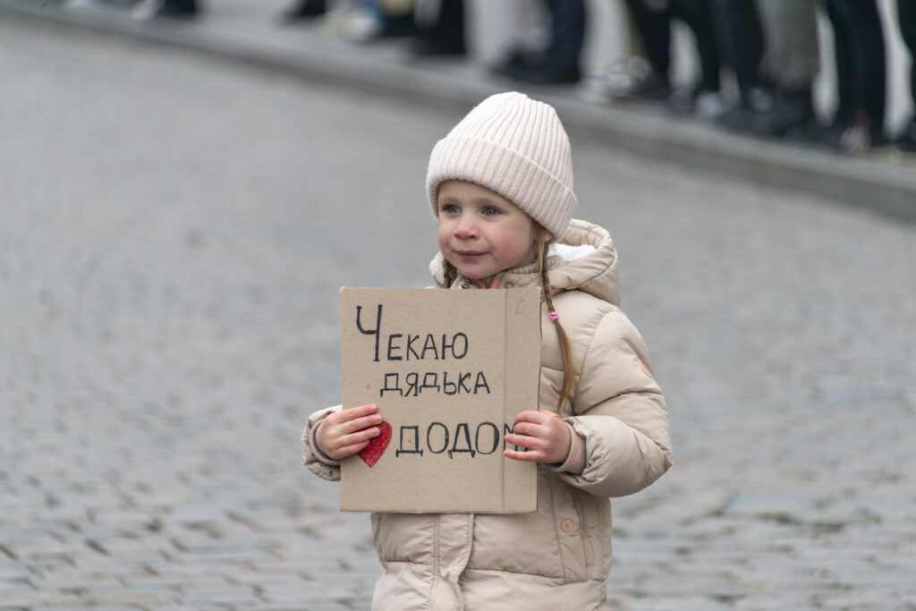 Ukraine – 23.03.2025: Ein kleines Mädchen steht im öffentlichen Raum im Zentrum von Odessa und hält ein handbeschriftetes Stück Karton vor sich. Darauf steht in kindlicher Schrift: „Ich warte auf Onkel Dodo.“ © Nataliia Mykhailenko / ukrainianphotodiary.org