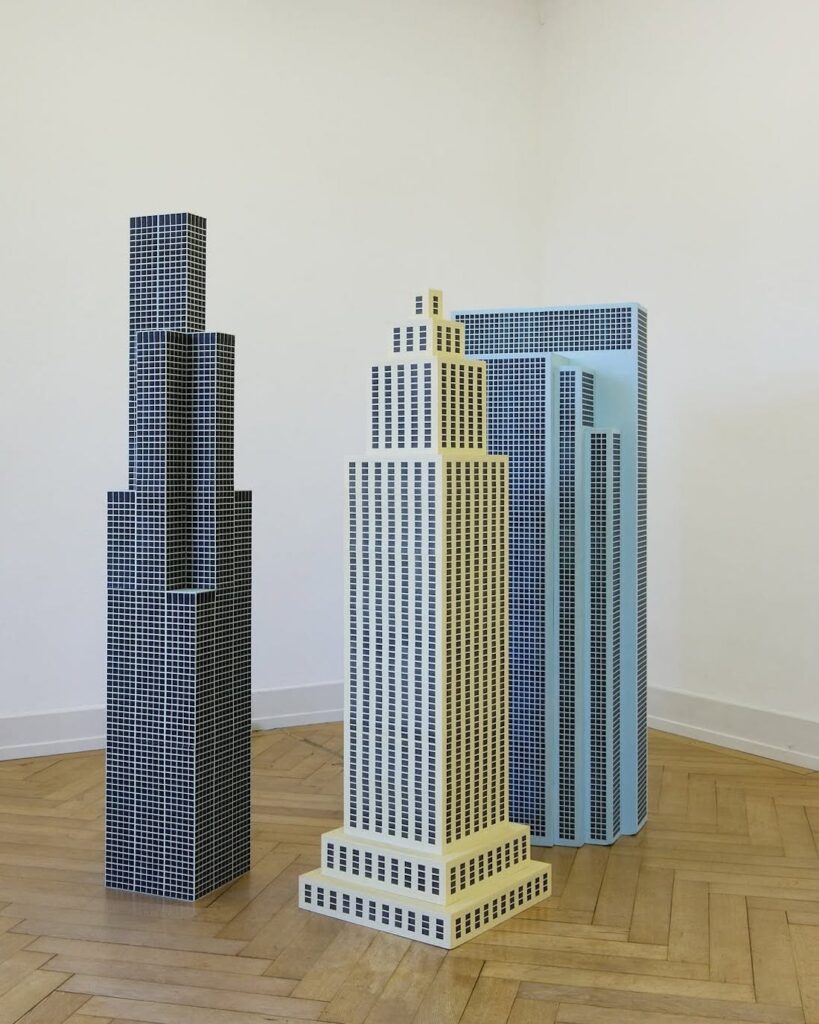 © Peter Brunner Brugg - Zu einer Dreiergruppe haben sich die «Das Mass aller Dinge» genannten Wolkenkratzer zusammengefunden. Jeweils in einer leicht unterschiedlichen Pastellfarbe gestaltet sind die Bauten von kleinen schwarzen Rechtecken und Quadraten geprägt, die sich wie ein Netz über die ganze Aussenseite legen. Die konstruktive, monumentale Wucht trifft auf feinste von Hand gemalte Spuren, die erst beim Anblick aus nächster Distanz sichtbar werden. Aus der Nähe offenbart sich auch, dass die Fenster zuerst gemalt und anschliessend mittels Fotokopie vervielfältigt und auf den Fassaden angebracht wurden.
